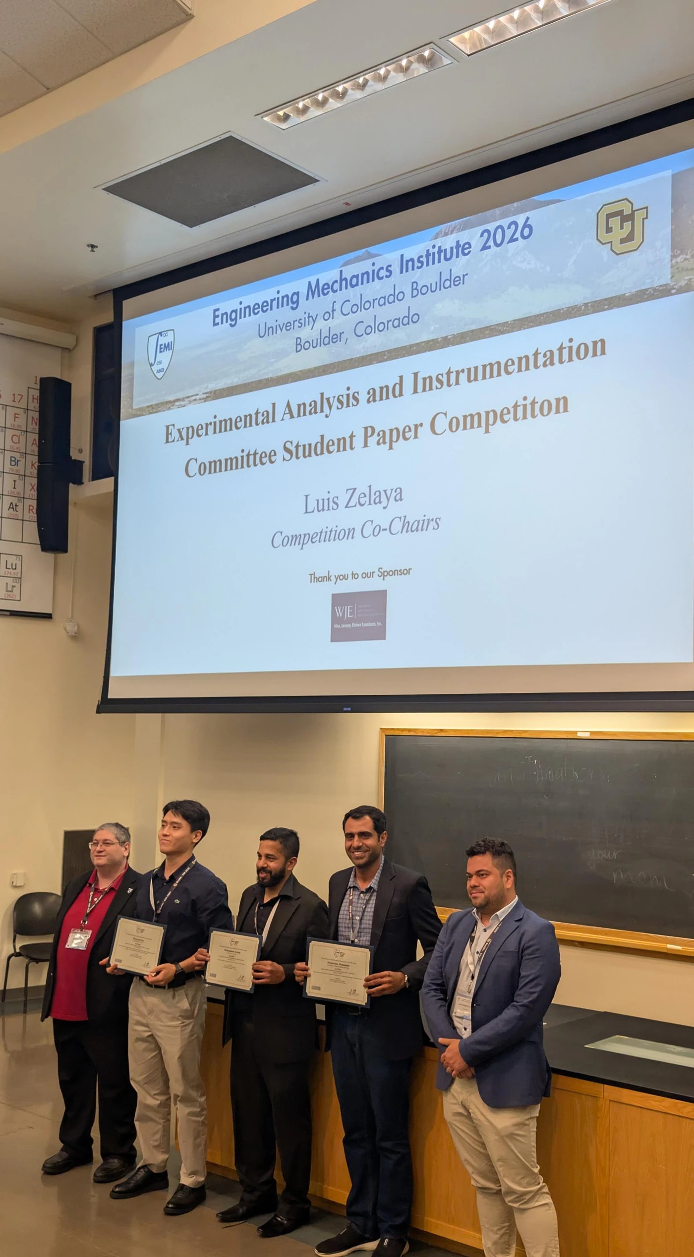 EMI 2026 award ceremony with certificate recipients in front of the projected competition recognition